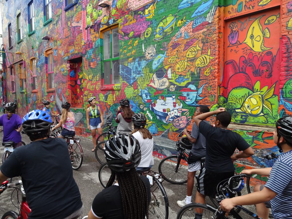 Group of students and adults bicycling in front of building with a colorful mural