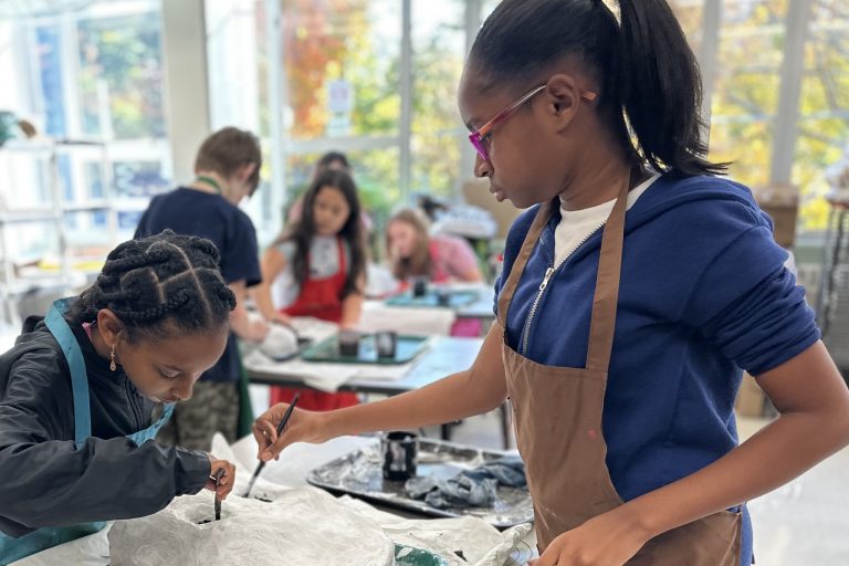 Two students in the foreground painting a Papier-mâché skull. More students are in the background doing the same.