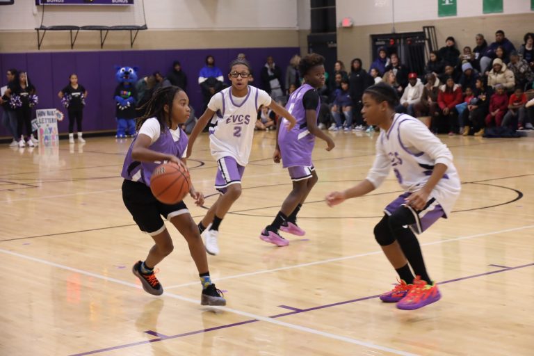 Four students playing a basketball game in jerseys, a crowd of cheerleaders and family are watching